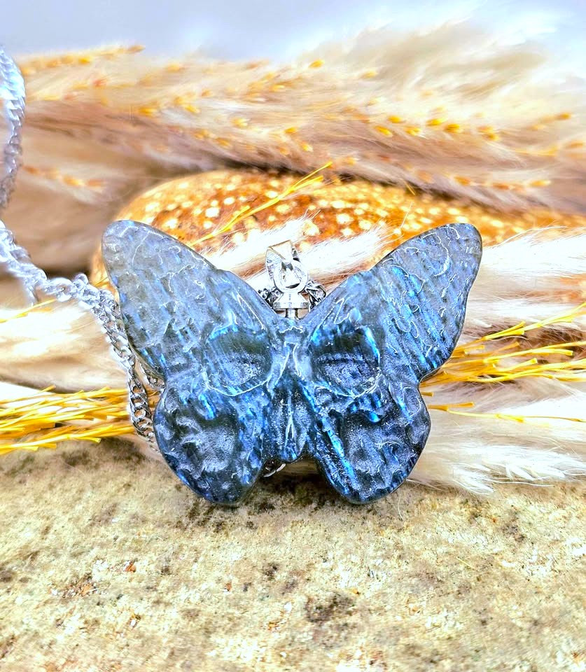 A blue iridescent butterfly with skull face at the center pendant necklace with a silver-toned metal clasp, displayed on a textured stone surface with dried yellow grass and soft beige feathers in the background.-My Aura Gemstones