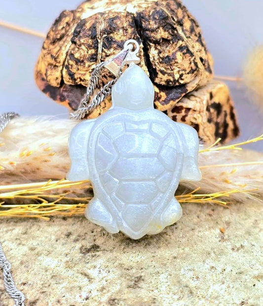 A carved turtle pendant necklace made of milky white moonstone, hanging from a silver chain, displayed on a textured stone surface with dried grass and a decorative rock in the background.-My Aura Gemstones