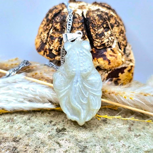 A moonstone carved goldfish pendant necklace with a silver chain, displayed on a textured stone surface with dried grass and a decorative object in the background.-My Aura Gemstones