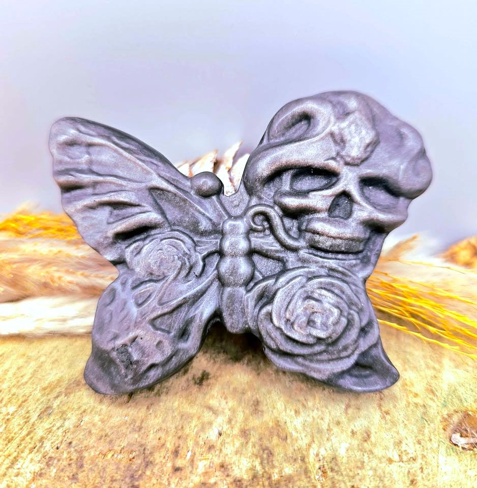 A dark gray gothic silver obsidian stone slice carved with butterfly with a skull on one wing and a rose on the other, resting on a textured surface with dried wheat.-My Aura Gemstones