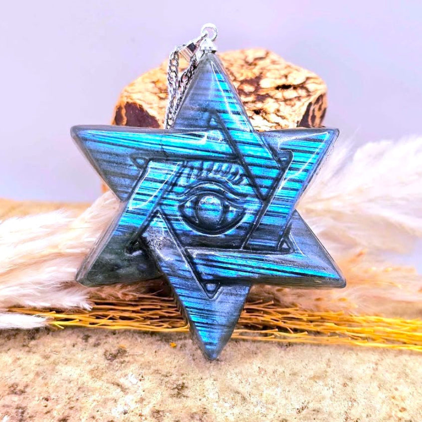 Natural Labradorite Star of David and all seeing eye pendant with an eye of providence symbol in the center, resting on a textured surface with dried grass and a small chain loop at the top-My Aura Gemstones