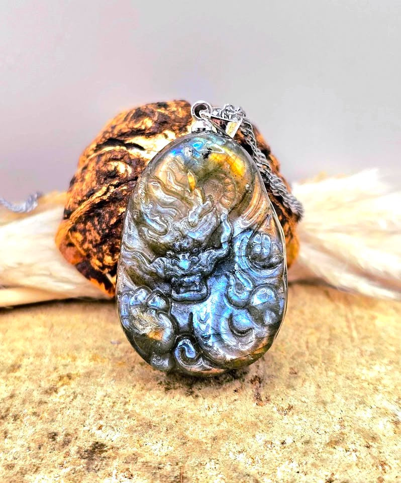 A teardrop-shaped labradorite pendant necklace featuring a detailed carved dragon design, resting on a textured brown shell and beige stone surface-My Aura Gemstones
