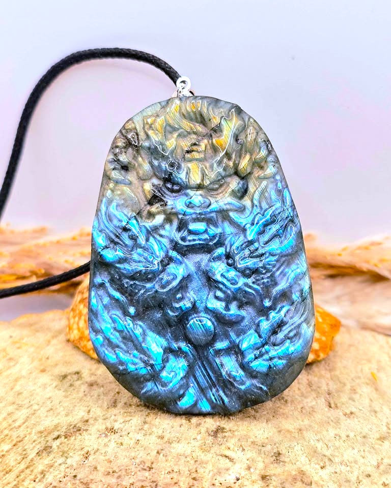 A carved stone pendant necklace featuring a detailed lion face design with a black cord, displayed on a sandy surface with a soft lavender background.-My Aura Gemstones