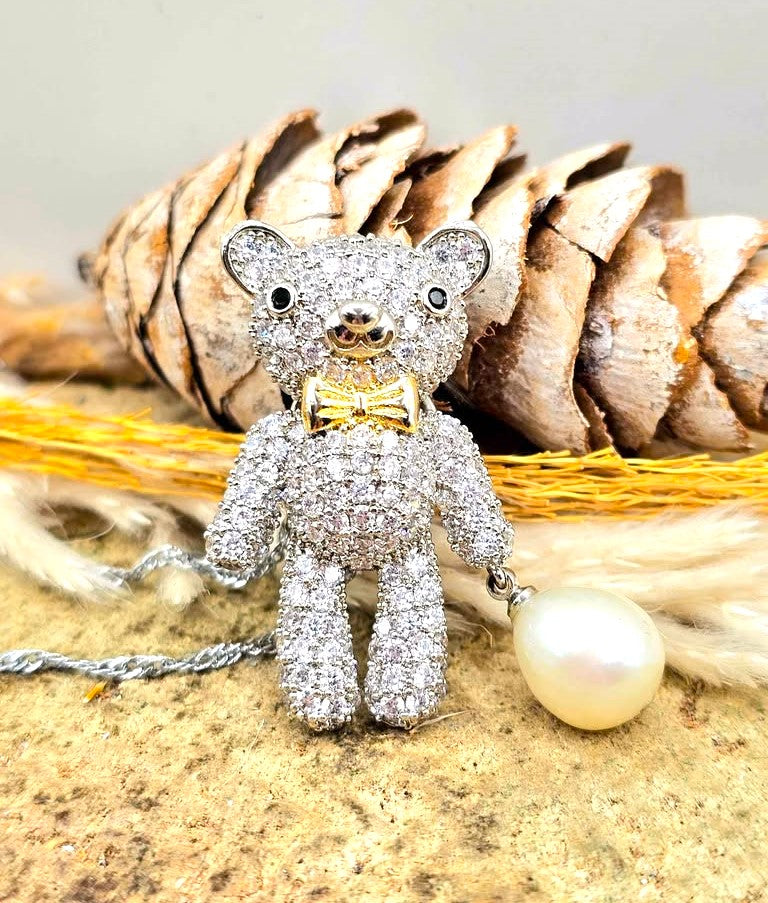 A teddy bear pendant covered in sparkling clear stones, wearing a gold bow tie, with a silver chain and a white pearl hanging from one hand-My Aura Gemstones
