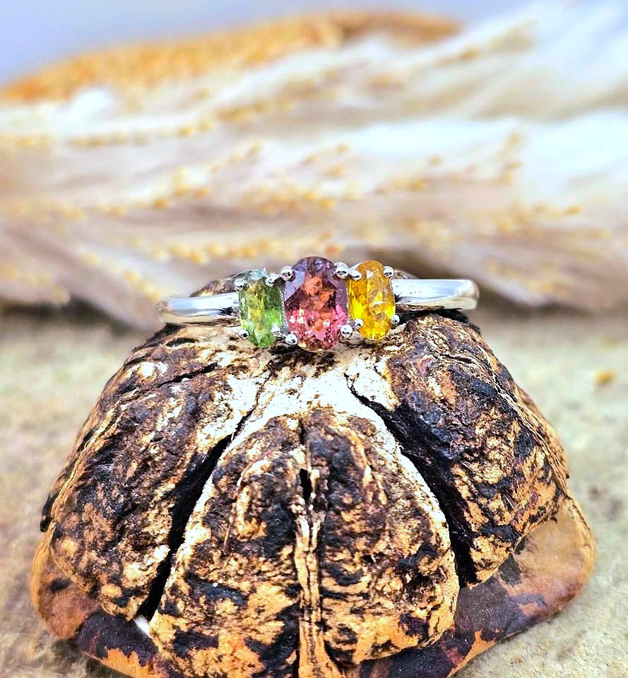A silver three-stone ring with green, pink, and yellow gemstones displayed on a textured brown shell against a sandy background.-My Aura Gemstones