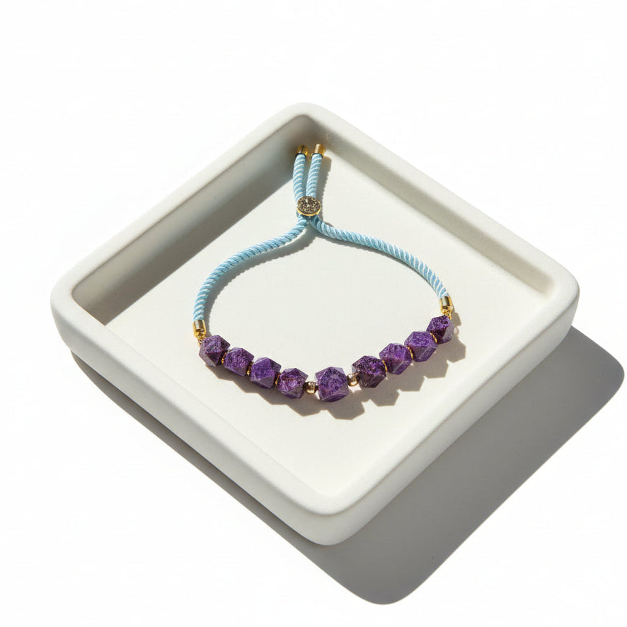 A light blue braided cord bracelet featuring faceted purple gemstone beads and gold-toned metal accents, displayed on a beige marble surface.
