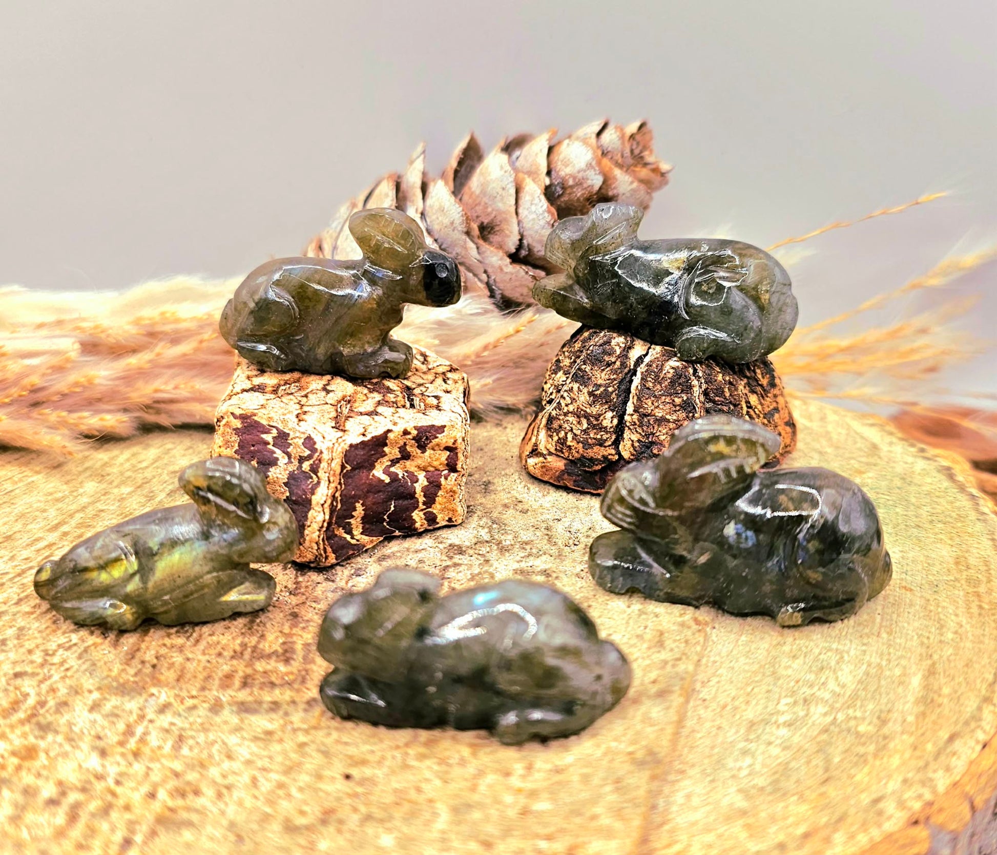Five polished nature labradorite  hand carved rabbit figurines displayed on a natural wood slice, with a pinecone and dried grass in the background.