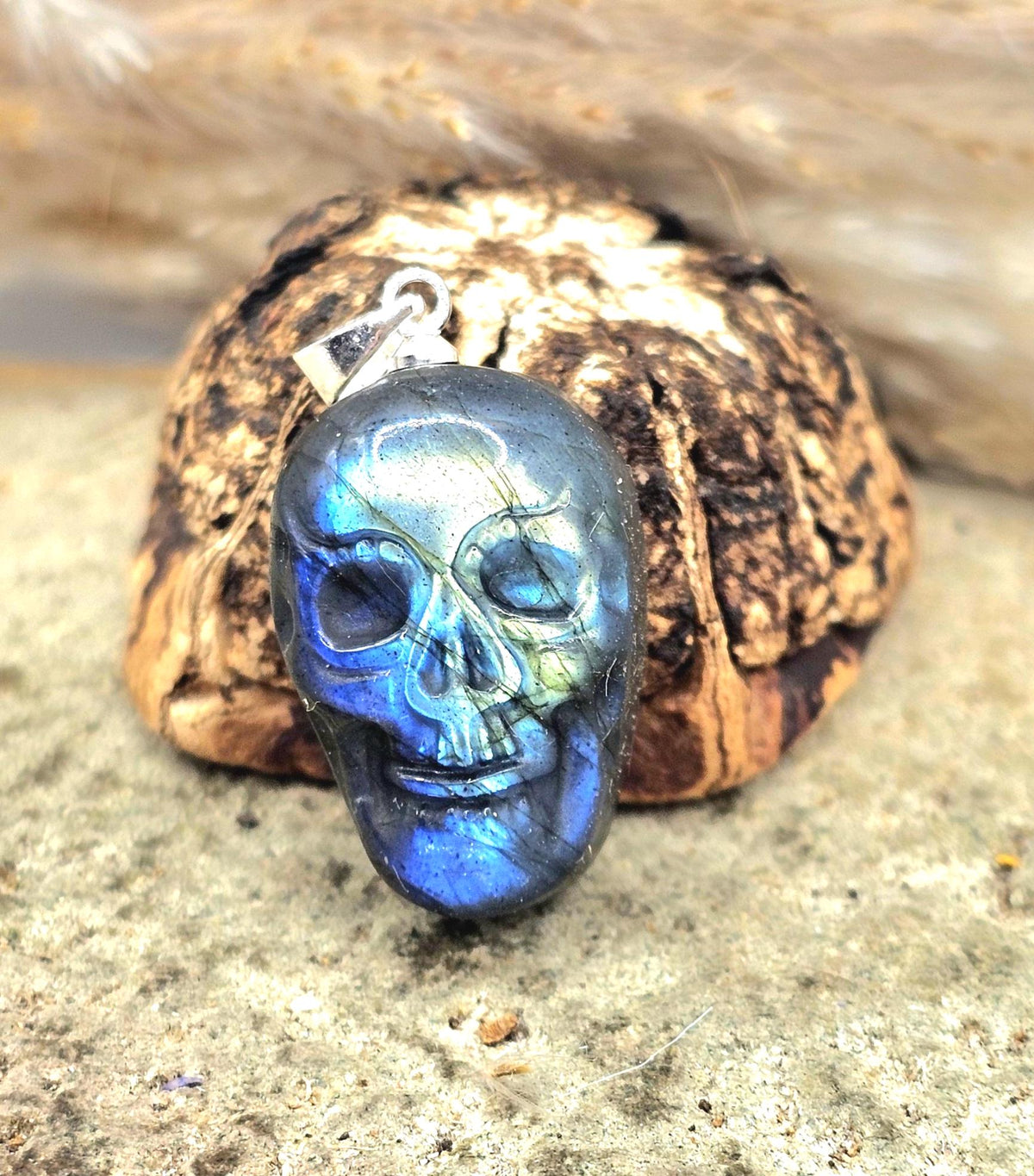 A polished skull-shaped pendant with a blue iridescent labradorite finish, shown resting on a piece of bark with a silver lobster clasp visible at the top, set against a natural stone background.