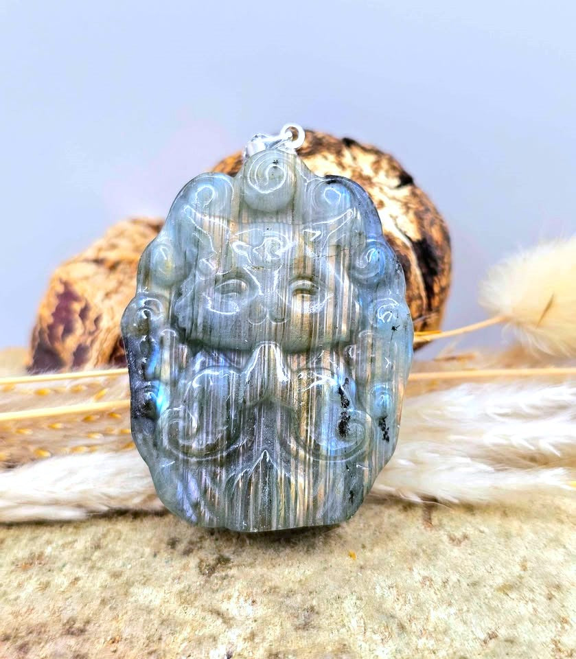 A blue labradorite pendant with a cat meditating on a lotus flower, resting on a textured surface with dried grass and a shell in the background. My Aura Gemstones